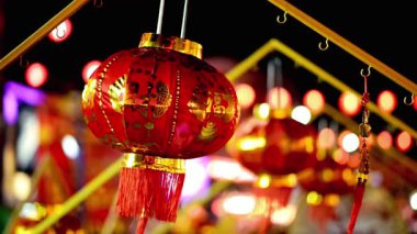 Göz kamaştırıcı kırmızı bir Çin feneri, altın aksanlı ve dalgalı püsküllerle süslenmiş canlı bir gece sahnesinde asılı duruyor. Kültürel bir kutlama ve şenlik neşesi hissi uyandırıyor..