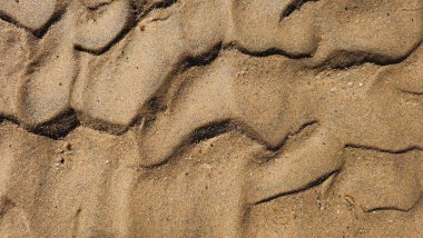 Kumsal kum dokusu. Geniş, dalgalı, kısa yatay çizgiler, dalgalanmalar ve rüzgarın oluşturduğu desenlerle dokulu bir kum yüzeyinin yakın çekimi. Renkler ışıktan koyu kahverengiye değişir.
