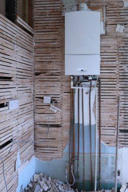 Bakır borular, elektrik prizleri ve yıkım enkazlarını gösteren, evin tadilatı sırasında lath ve alçıyla çevrili duvara monte edilmiş bir kazan. Endüstriyel, dağınık yenileme sahnesi onarım ve onarım işleri.