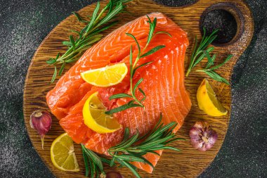 Somon balığı. Taze çiğ somon balığı filetosu. Pişirme malzemeleri, otlar ve limon dilimleme tahtasında, siyah yeşil arka plan, üst görünüm. 