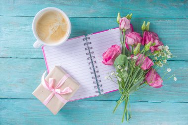 Yaz sabahı tatil arkaplanı, tebrik ya da davetiyeler için. Mesajın için not defteri, Kraft kağıdında hediye kutusu, bir fincan sabah kahvesi ve mavi ahşap arka planda bir buket çiçek.