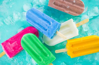 Selection of bright multicolored ice cream popsicle. Various gelato, frozen lollypops - chocolate vanilla blueberry strawberry pistachio orange, with crushed ice on light blue sunny background clipart