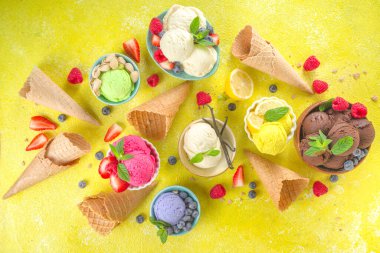 Farklı kaselerde çeşitli renkli dondurma topları, dondurma külahları ve aromalı malzemeler - şam fıstığı, böğürtlen, limon, çikolata, vanilya, nane. Renkli sarı parlak arkaplan