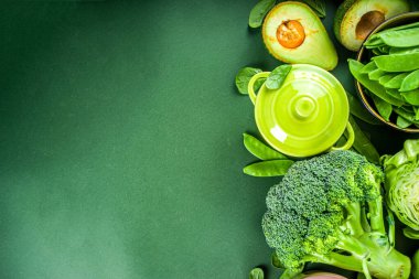 Koyu yeşil mutfak masasında içi boş porsiyonlu tencerelerle çeşitli organik yeşil bahar sebze malzemeleri, üst manzara. Diyet sağlıklı çorba, güveç yemek hazırlama konsepti