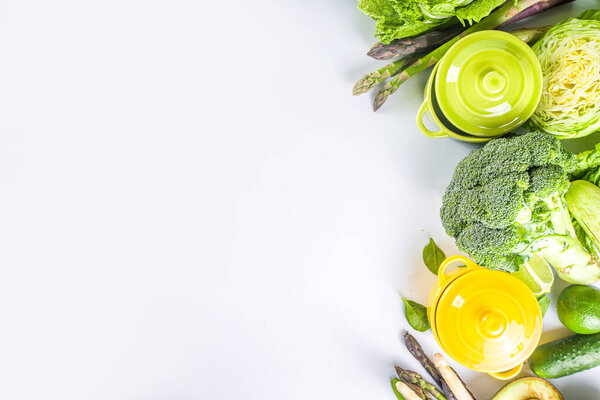 Various organic green spring vegetable ingredients with empty colorful portioned cooking pots on white kitchen table, top view. Diet healthy soup, stew food preparation concept