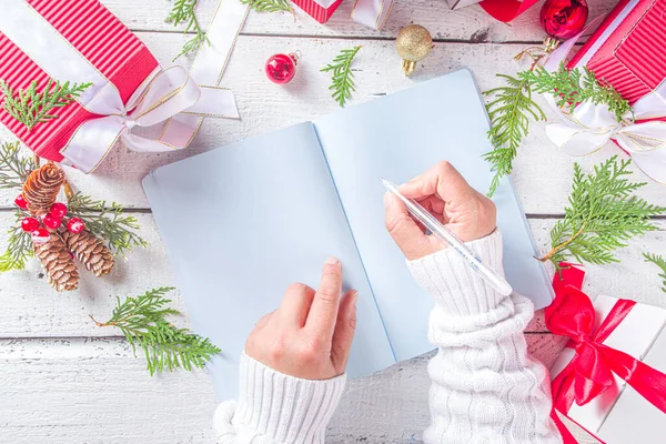 Not defterine noel süsü ve beyaz ahşap masada hediye kutuları olan bir kadın el yazısı. Noel dilek listesi, Noel Baba mektubu, liste konsepti, düz kopya alanı