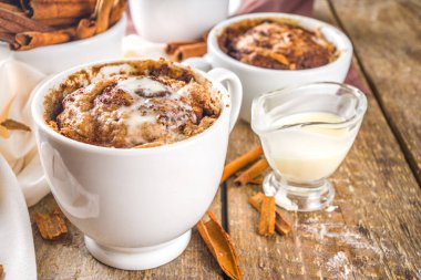 Kupada tarçınlı kek. Basit bir mikrodalga tatlı fikri, tarif için arka plan. Tarçınlı kek, üzerine şeker ve krem peynir konmuş, farklı bardaklarda, ahşap arka planda tarçın çubukları ile