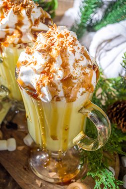 Ev yapımı Tuzlu Karamelli Eggnog veya Beyaz Sıcak Çikolatalı içecek, karamel soslu, krem şantili ve marşmelovlu, ahşap arka planda, Noel süslemeleri ile birlikte