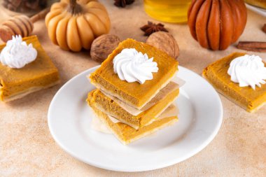 Homemade autumn taste pumpkin pie bars. Freshly baked pumpkin bars with whipped cream on it, with autumn decor and spices on beige table background, copy space 