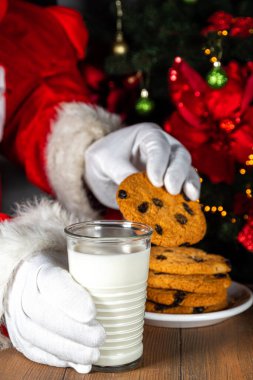 Noel Baba kurabiye ve süt yiyor. Noel Baba elleri Noel ağacına karşı ahşap masada kurabiye ve süt bardağı topluyor., 
