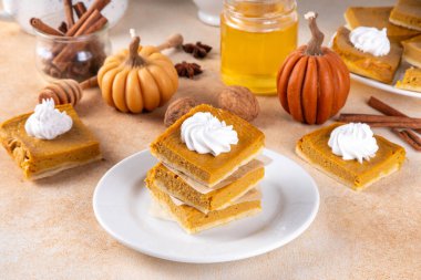 Homemade autumn taste pumpkin pie bars. Freshly baked pumpkin bars with whipped cream on it, with autumn decor and spices on beige table background, copy space 
