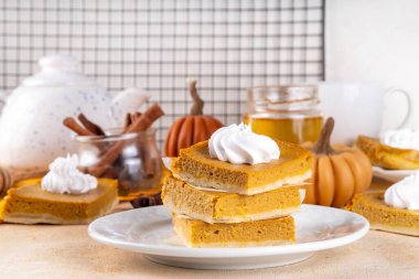 Homemade autumn taste pumpkin pie bars. Freshly baked pumpkin bars with whipped cream on it, with autumn decor and spices on beige table background, copy space 