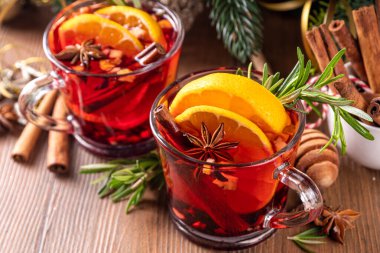 Geleneksel Noel aromalı şarap kokteyli, baharatlı kırmızı şarap içkisi, tarçın, portakal ve biberiye filizleri, Noel süslenmiş ahşap kaplama arka planda.