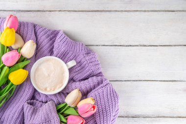 Kahve arkaplanlı bir bahar sabahı. Bir fincan kahve latte, leylak kadın örgü süveter, bir buket lale çiçeği. Bahar tatili, tatil arkaplanı, tebrik kartı, üst görünüm düz kopya alanı