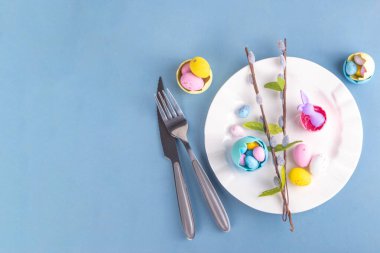 Paskalya Bayramı menü seti, pastel mavi arka planda çatal bıçak takımı ve tabak kareli peçete, şenlikli kurdele, şirin tavşanlı Paskalya bayramı dekoru, paskalya çikolatası yumurtaları.