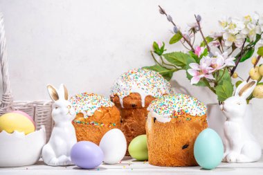Şekerli Paskalya pastaları, renkli yumurta sepeti, seramik tavşanlar ve beyaz mutfak masasında bahar çiçekleri, fotokopi alanı. Mutlu Paskalya Tatilleri tebrik kartı arkaplan kopyalama alanı