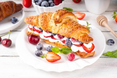 Krem şantili, taze meyve ve meyveli tatlı kahvaltı kruvasan sandviçi. Lezzetli bir yaz yemeği ya da içinde çilek, yabanmersini, kiraz, ahududu ve fındık bulunan taze kruvasanla atıştırmalık bir şeyler.