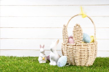 Paskalya yumurtası avcılığı geçmişi. Paskalya sepetinde renkli Paskalya yumurtası, çikolatalı yumurta, tavşan ve tavşan dekoru yeşil çimen bahçesi arka planında beyaz ahşap duvarlı