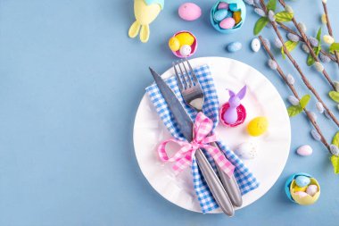 Paskalya Bayramı menü seti, pastel mavi arka planda çatal bıçak takımı ve tabak kareli peçete, şenlikli kurdele, şirin tavşanlı Paskalya bayramı dekoru, paskalya çikolatası yumurtaları.
