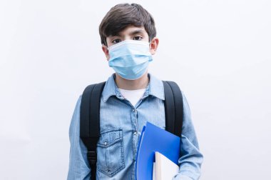 Sırt çantalı ve maskeli güzel çocuk beyaz arka planda kitap tutuyor.