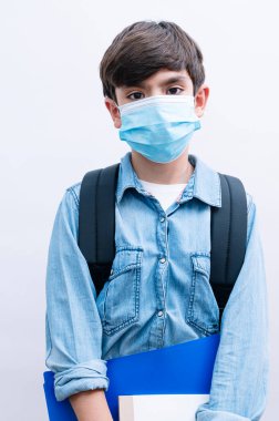 Sırt çantalı ve maskeli güzel çocuk beyaz arka planda kitap tutuyor.