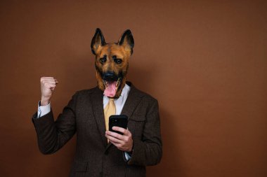 Akıllı telefon ve lateks köpek kafası maskeli neşeli bir adam ellerini kaldırıyor.