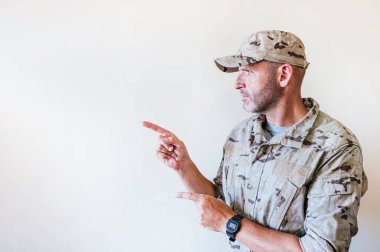 Kamuflaj üniformalı yakışıklı adam gülümsüyor ve iki eli ve parmakları yanda işaret ediyor..
