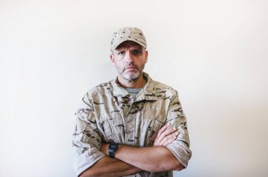 Kamuflajlı ordu üniformalı beyaz adam kollarını kavuşturuyor.