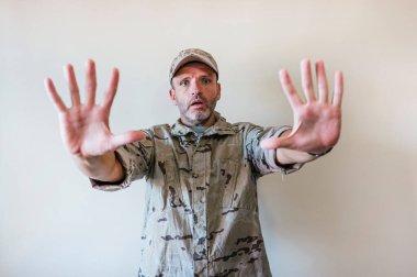 Kamuflajlı asker üniforması giymiş beyaz bir adam elinde el hareketi, öfke ve hüsranın ifadesi ile durduruyor.