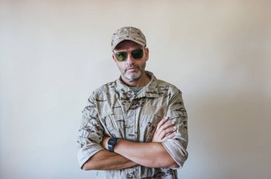 Kamuflajlı ordu üniformalı beyaz adam kollarını kavuşturuyor.