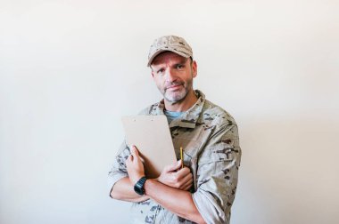Caucasian man in camouflaged military uniform holding notebook.