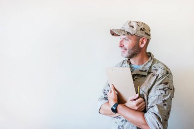 Caucasian man in camouflaged military uniform holding notebook.