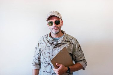 Caucasian man in camouflaged military uniform holding notebook.