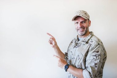 Kamuflaj üniformalı yakışıklı adam gülümsüyor ve kameraya bakıyor. İki eli ve parmağı yana bakıyor..