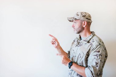 Kamuflaj üniformalı yakışıklı adam gülümsüyor ve iki eli ve parmakları yanda işaret ediyor..