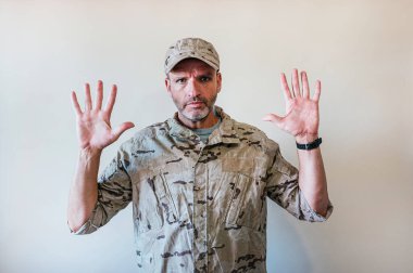 Kamuflajlı asker üniforması giymiş beyaz bir adam elinde el hareketi, öfke ve hüsranın ifadesi ile durduruyor.