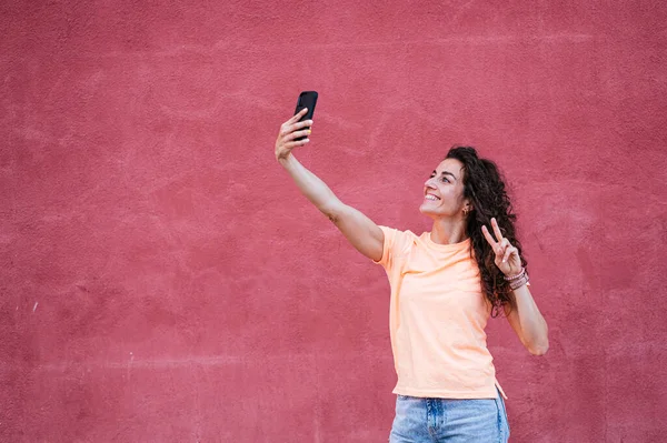 Mulher Tomando Selfie Smartphone Enquanto Mostra Sinal Paz Sorrindo — Fotografia de Stock