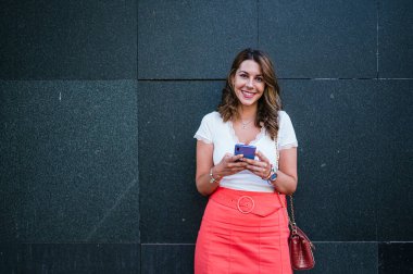 Yazlık elbiseli bir kadın akıllı telefon kullanıp şehirde dikilirken görülüyor..