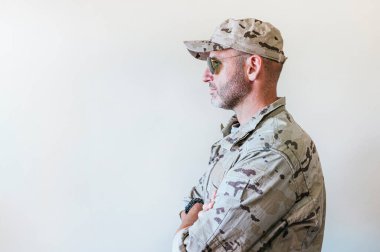 Kamuflajlı ordu üniformalı beyaz adam kollarını kavuşturuyor.