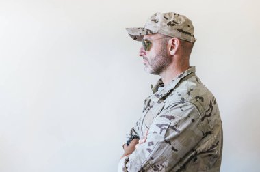 Kamuflajlı ordu üniformalı beyaz adam kollarını kavuşturuyor.