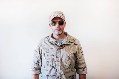 Kamuflajlı ordu üniformalı beyaz adam kollarını kavuşturuyor.