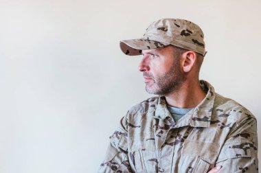 Kamuflajlı ordu üniformalı beyaz adam kollarını kavuşturuyor.