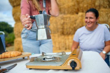 Bir kadın dışarıda gülümserken portatif sobanın yanında kahve demliği tutan bir kadın..