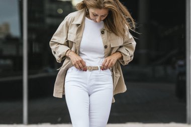 Bej ceketli ve beyaz pantolonlu kadın kemerini ayarlıyor..