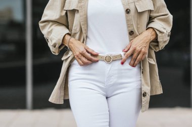 Bej ceketli ve beyaz pantolonlu kadın kemerini ayarlıyor..
