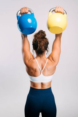 Kadın, mavi ve sarı çaydanlık çanlarıyla üstten baskı yapıyor, güçlü sırt kaslarını gösteriyor.