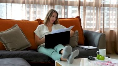 Şirin bir kadın kağıt dökümanlara bakar, kanepeye ya da kanepeye dizüstü bilgisayarla oturur.