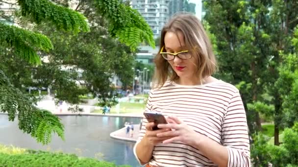 Cute młoda kobieta używa czarny smartfon na zielonym tle drzew — Wideo stockowe
