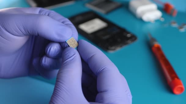 Mavi eldivenler parçalanmış akıllı telefon arkaplanındaki mikro SIM kartı tutuyor. — Stok video