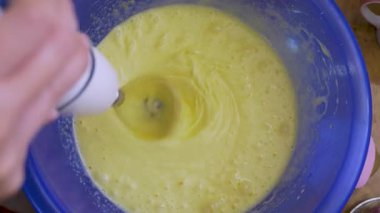 Mavi kasenin üstündeki el çırpma makinesini kadın kontrol ediyor. Yumurta sıvısı köpükler hafif. Mutfak çırpıcı krep hazırlama sırasında ev mutfağında aktif olarak hareket eder